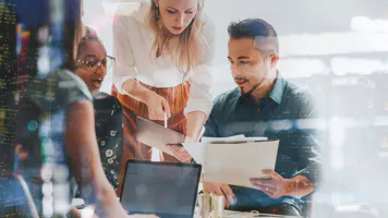 A diverse group of four colleagues collaborate around a desk with laptops and documents. Overlaid cityscape reflects a dynamic, innovative workplace ambiance.